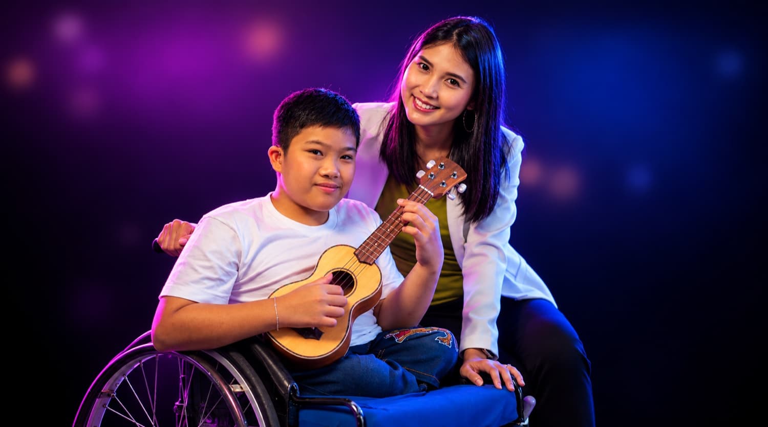 A smiling boy in a wheelchair holds a ukulele while a young woman leans in beside him, against a purple and blue stage-lit background